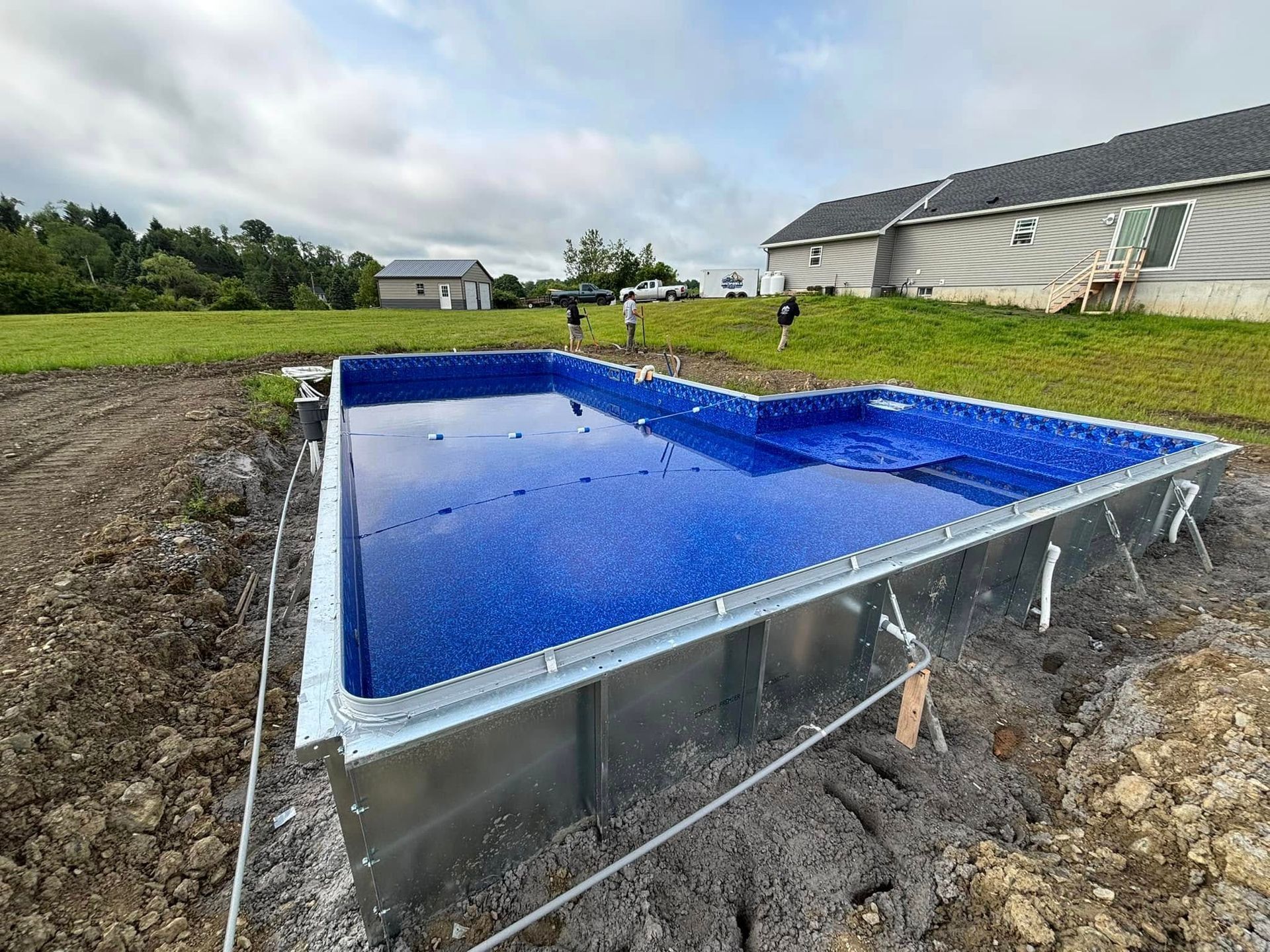 Partially constructed rectangular swimming pool, blue interior, in a backyard. Workers in the background. Cloudy sky.