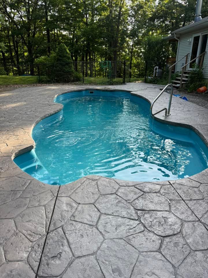 Swimming pool with blue water surrounded by textured concrete patio. Trees and house in the background.