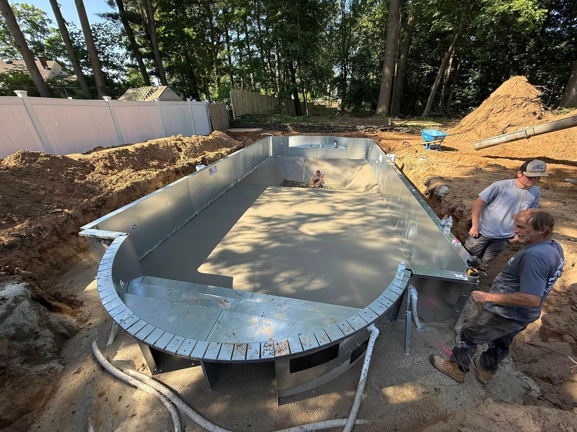 Two men work on a concrete pool under construction. Steel frame, dirt, and trees surround the pool.