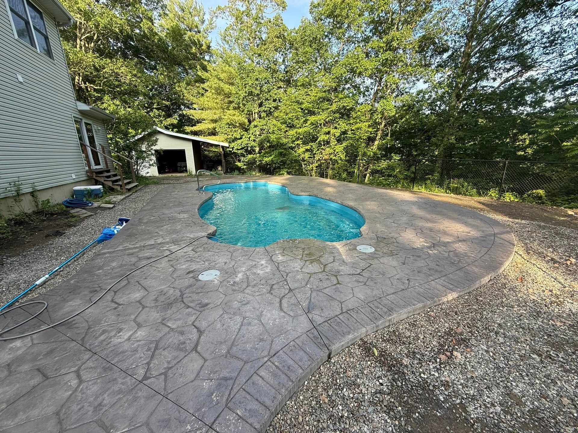 Pool with textured concrete patio, set in a backyard. House and small shed visible, surrounded by trees.