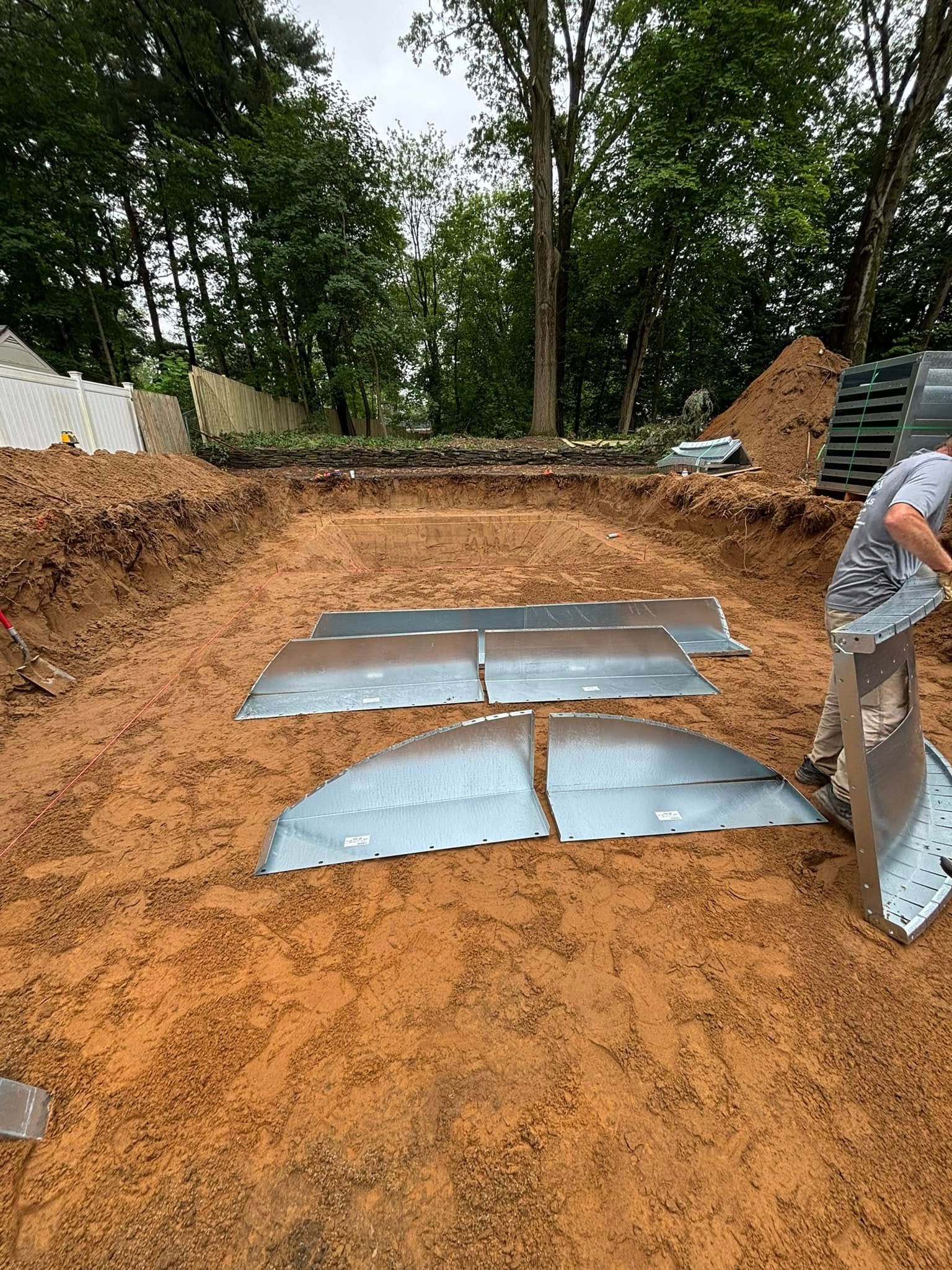 A construction site: a man assembling metal panels in a dug-out area, with trees in the background.