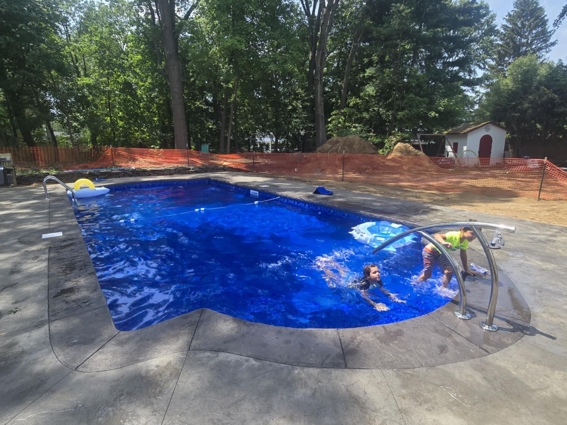People swimming in a blue-tiled pool with concrete decking; trees and a small shed in the background.