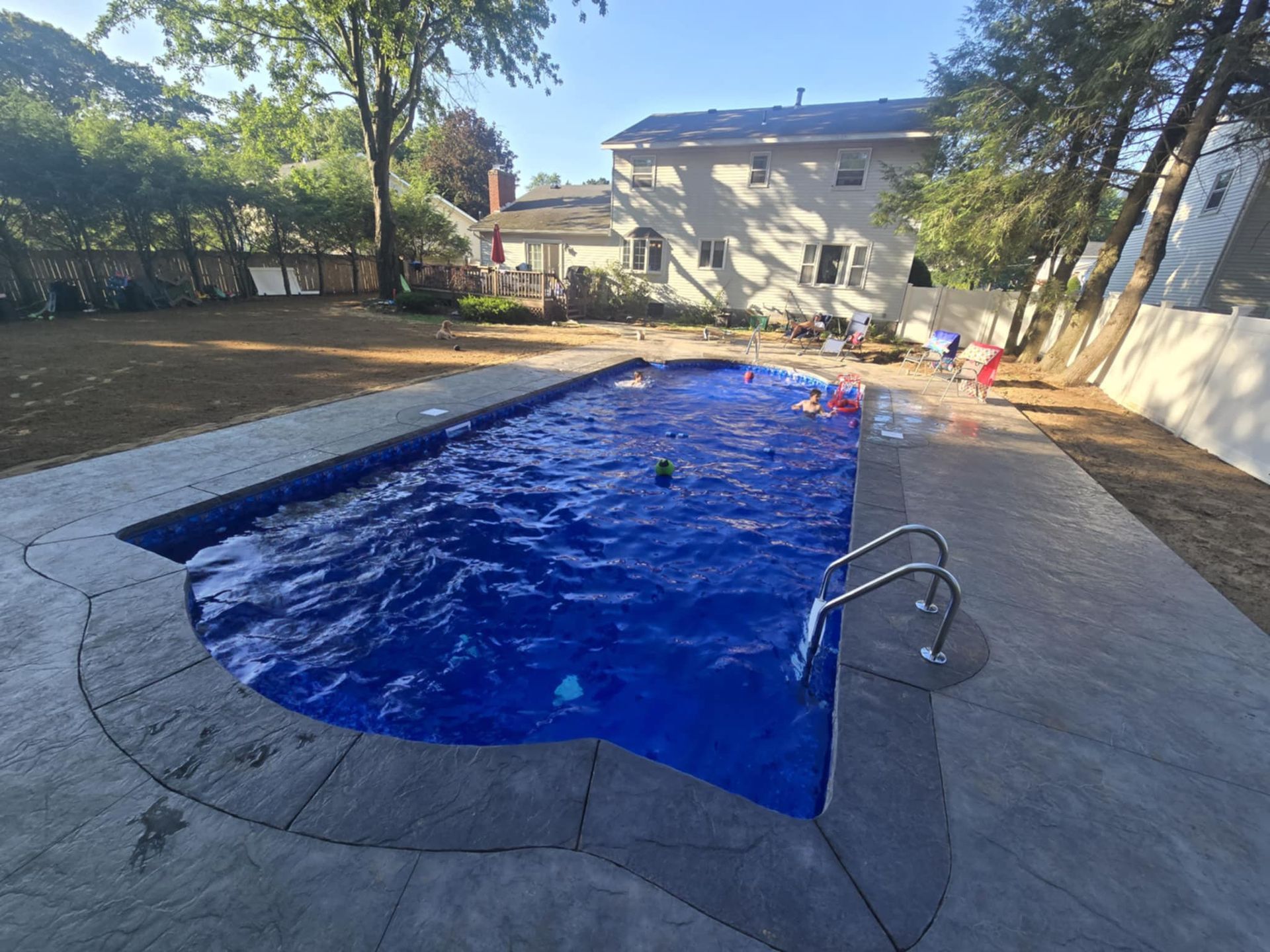 Backyard with a blue swimming pool. People swim in the pool with surrounding concrete. Green grass and trees visible.