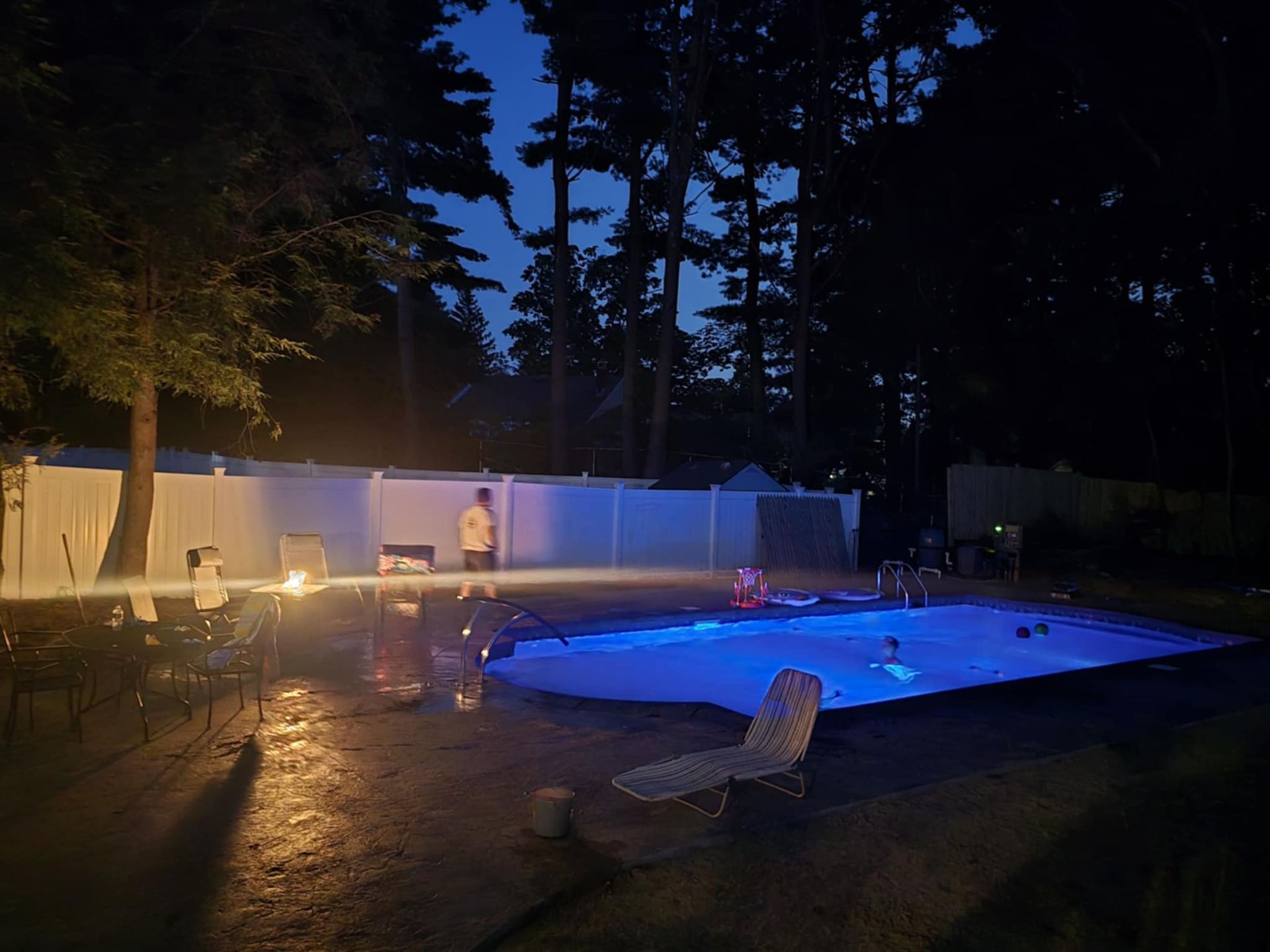 Pool at night, lit by blue lights, with people swimming. Trees and white fence in the background.