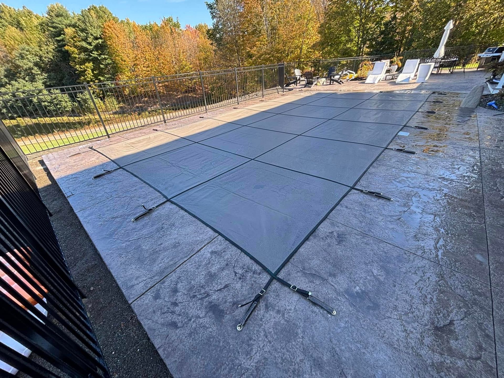 Pool covered with a gray mesh safety cover on a concrete patio, surrounded by trees and a black fence.