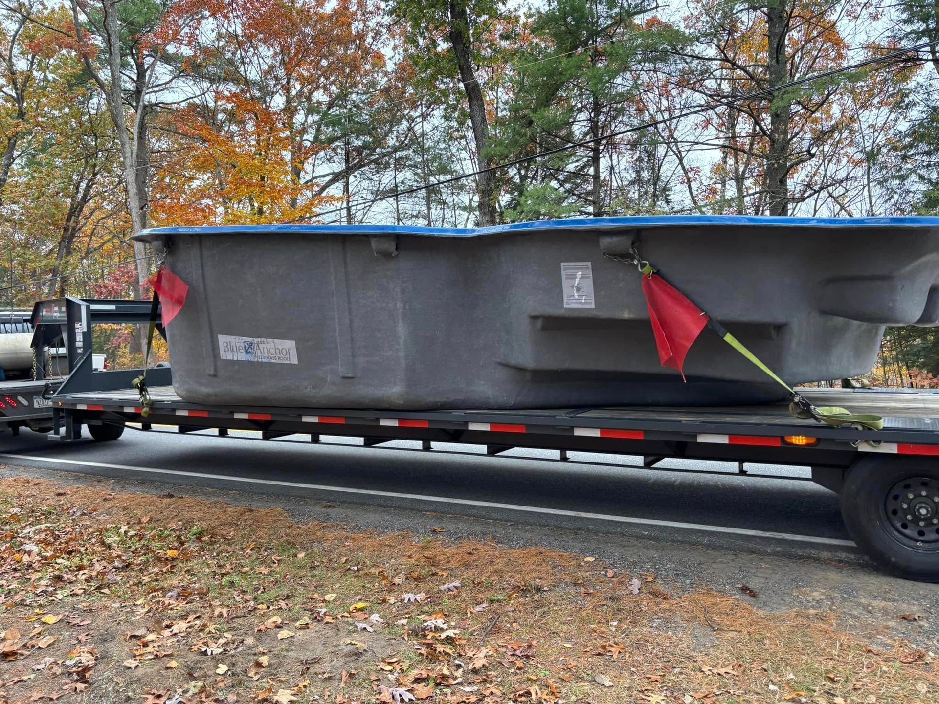 A large gray rectangular pool being transported on a flatbed trailer on a road with fall foliage.