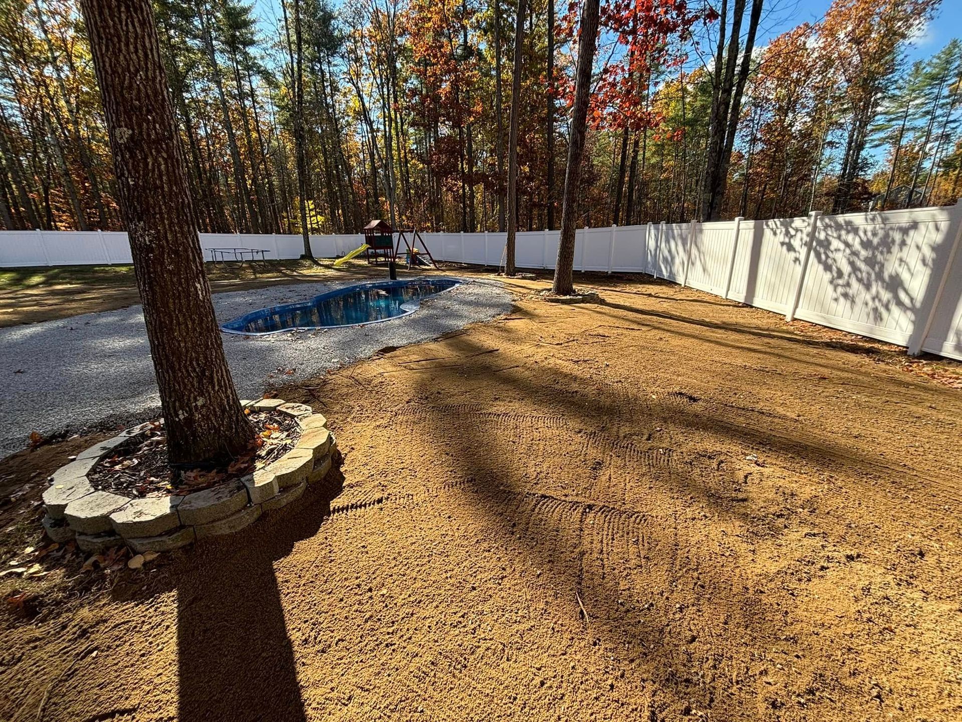 Backyard with gravel path, small pool, trees, and white fence in autumn.