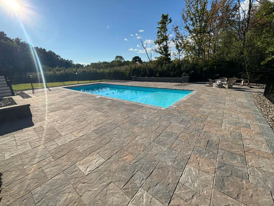 A rectangular pool surrounded by a stone patio, with a bright sky and trees in the background.