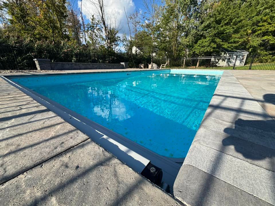Blue swimming pool with stone coping and surrounding patio, trees, and sky.
