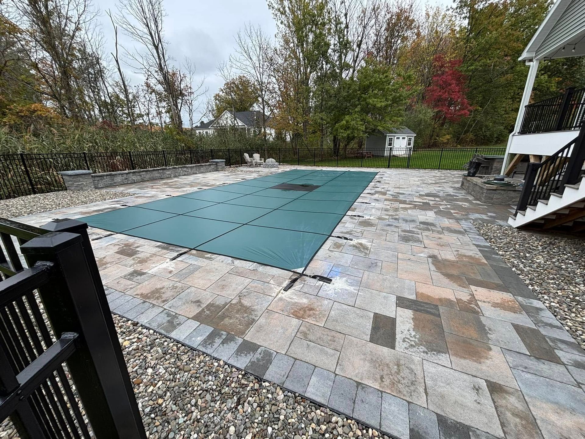 Pool covered with a green tarp, surrounded by paved patio. Black fence and steps on the right.