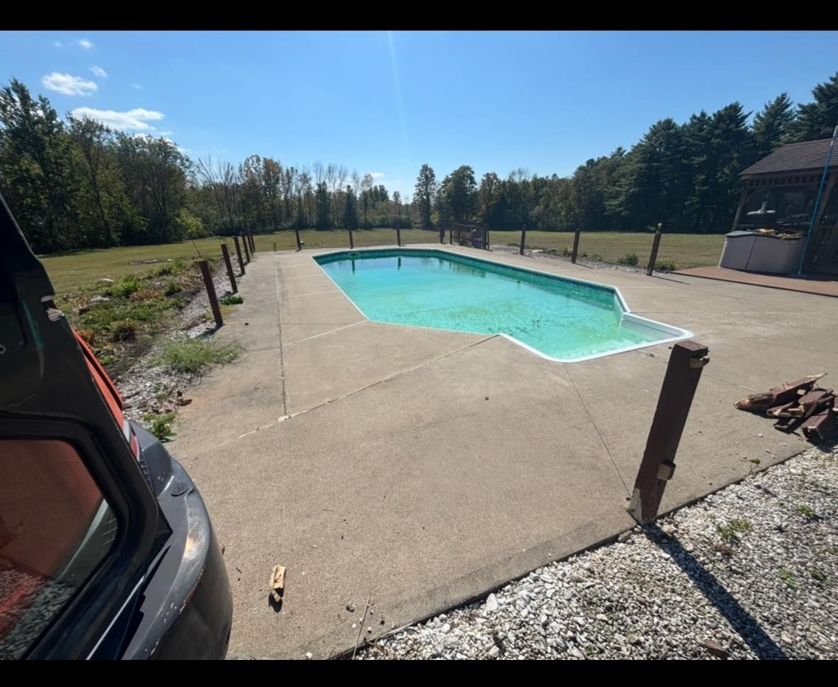 An empty, algae-covered outdoor pool surrounded by concrete, a grassy area, and trees on a sunny day.