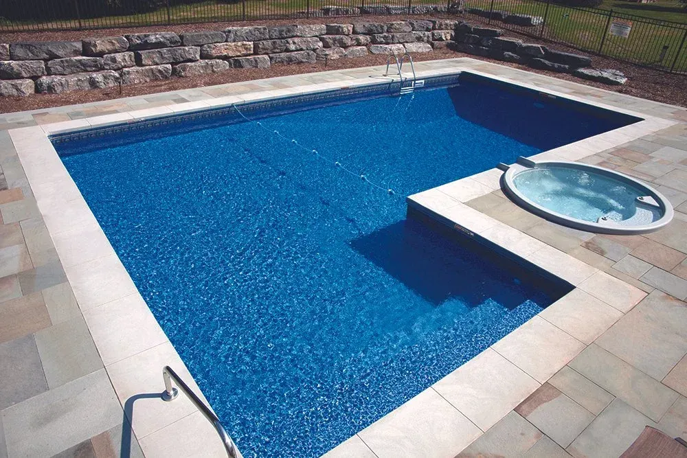 Swimming pool with built-in spa, concrete patio, steps. Blue water and a stone retaining wall.