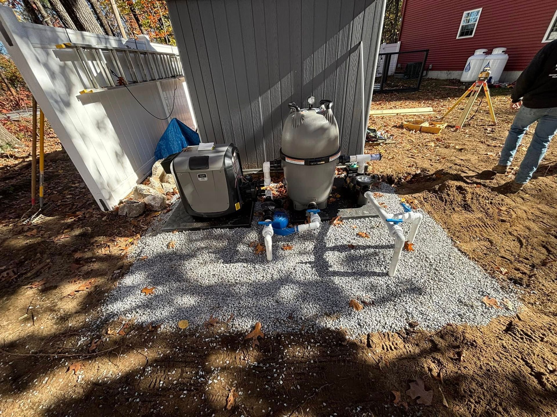 Pool equipment setup on gravel, with pump, filter, and plumbing. A person stands nearby.