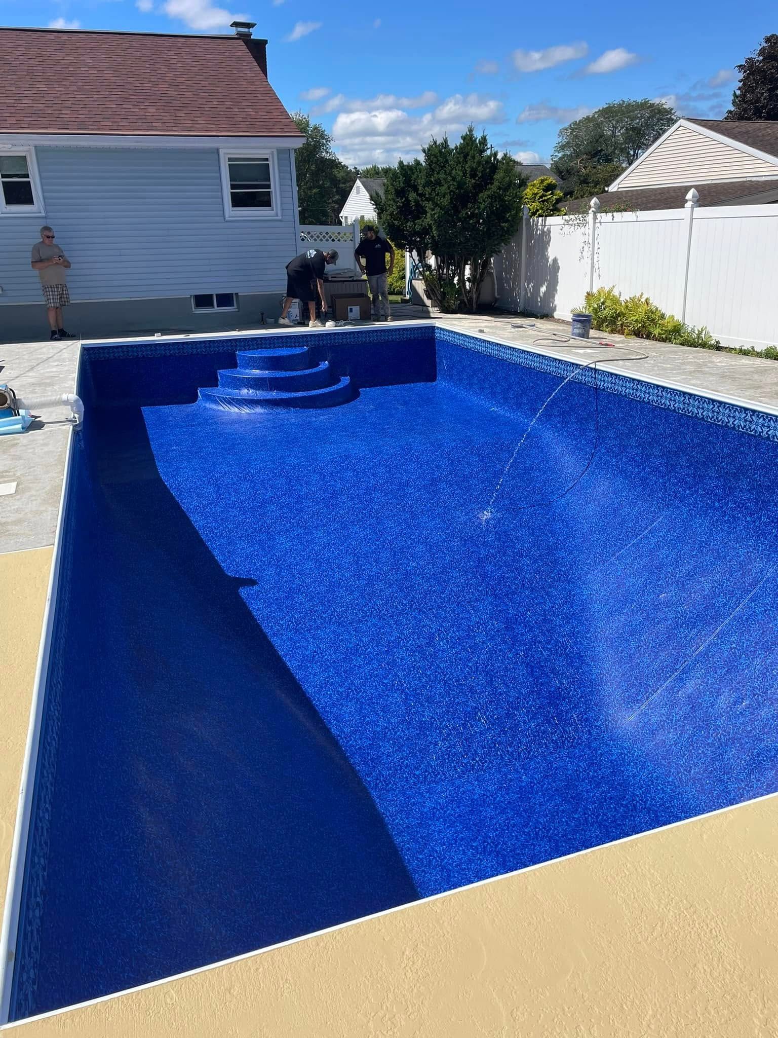 Empty in-ground pool with blue tile interior. Workers near the edge, house and white fence in background. Sunny day.