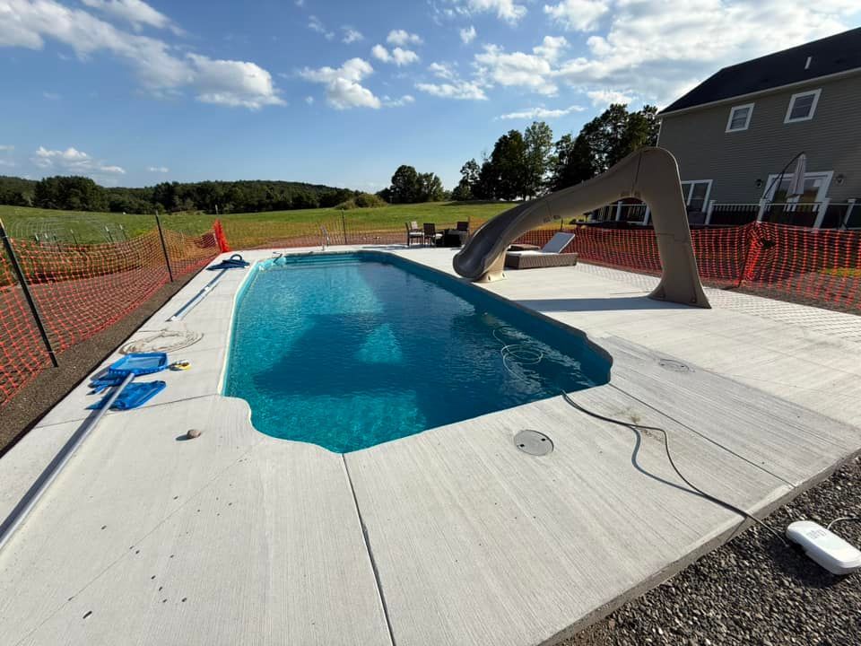 Swimming pool with a water slide surrounded by concrete, in front of a house under a sunny sky.