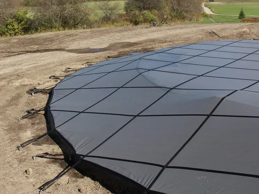 Gray pool safety cover stretched across a dirt area with tie-down straps.
