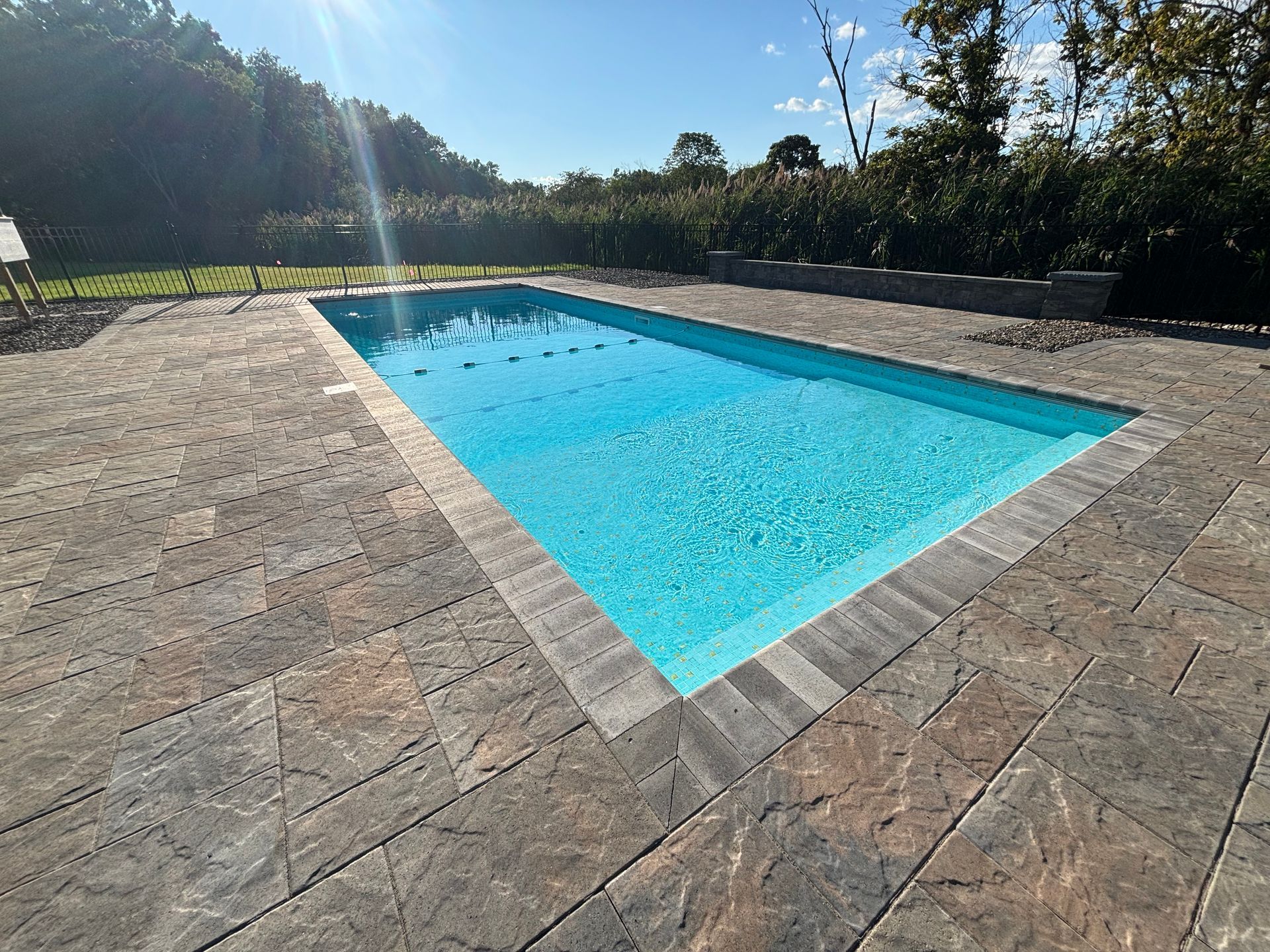 Rectangular teal pool with white trim, surrounded by gravel, next to white and black buildings.