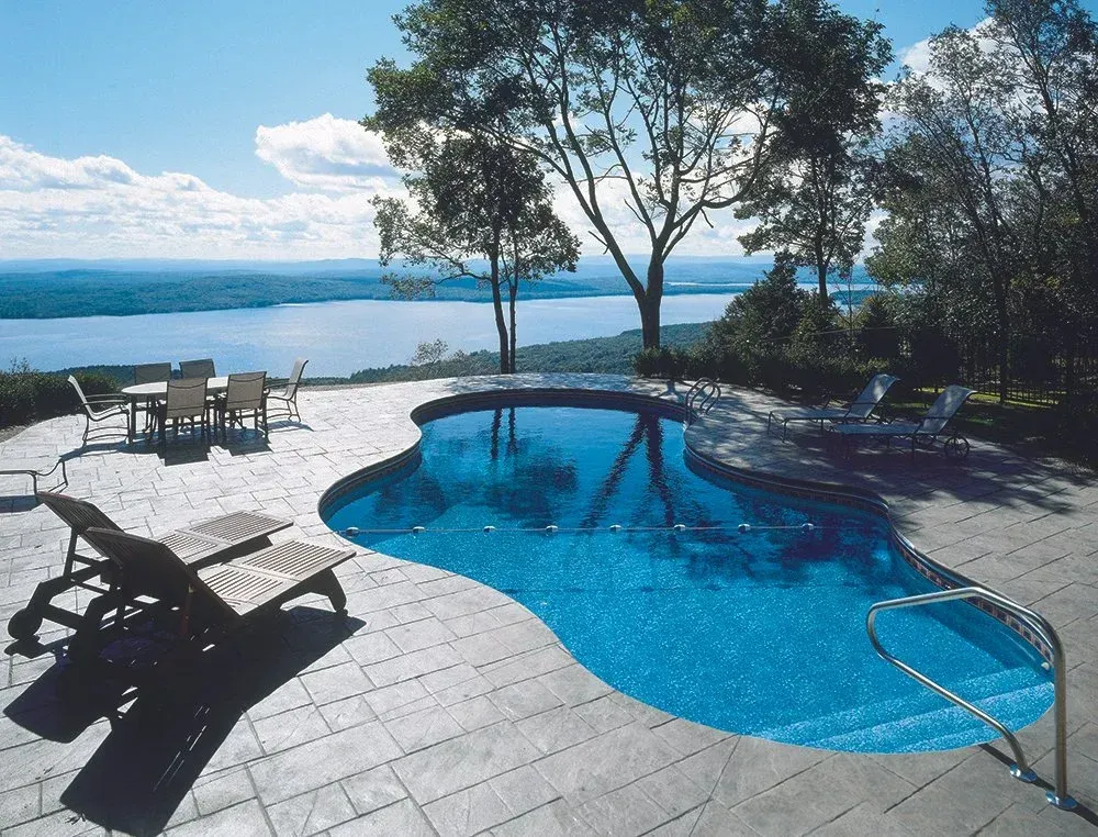 Pool overlooking lake; lounge chairs, dining table on patio. Trees, blue sky, and water.