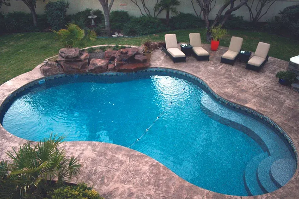 Pool with stone waterfall, tanning chairs on stone patio, green lawn in background.