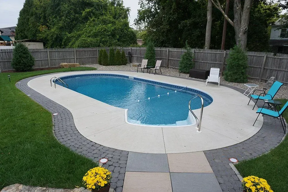 Backyard pool with paved patio, grass, and surrounding fence.