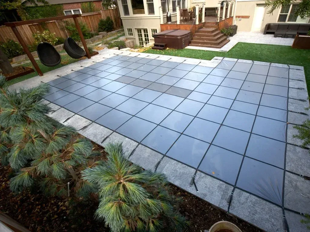 Pool covered with a gray safety cover in a backyard with landscaping, swings, and a deck.