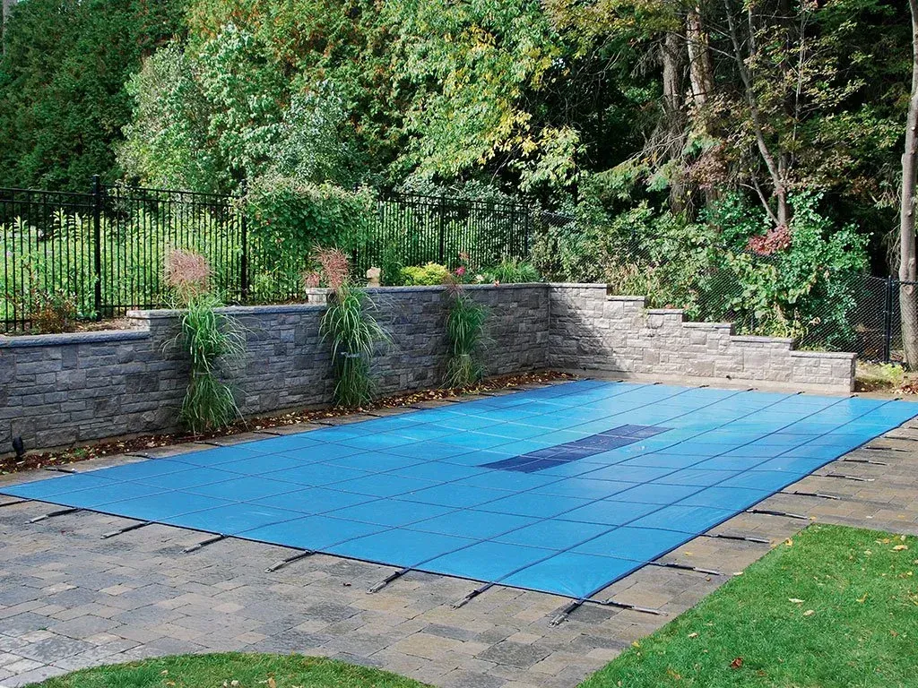 Blue pool cover over a rectangular pool in a backyard with stone wall and foliage.