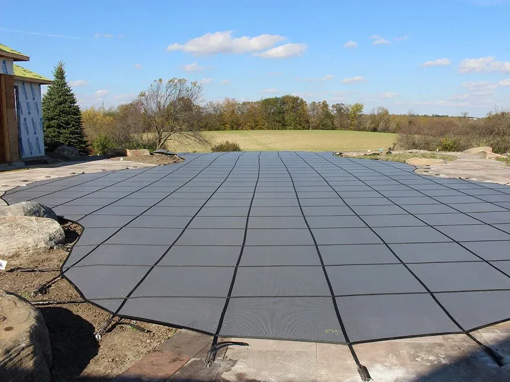 A gray pool cover on a concrete patio, with a grassy landscape and blue sky in the background.