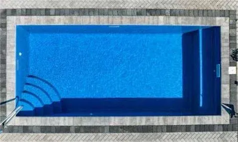 Overhead view of a rectangular blue swimming pool with steps, surrounded by gray paving stones.