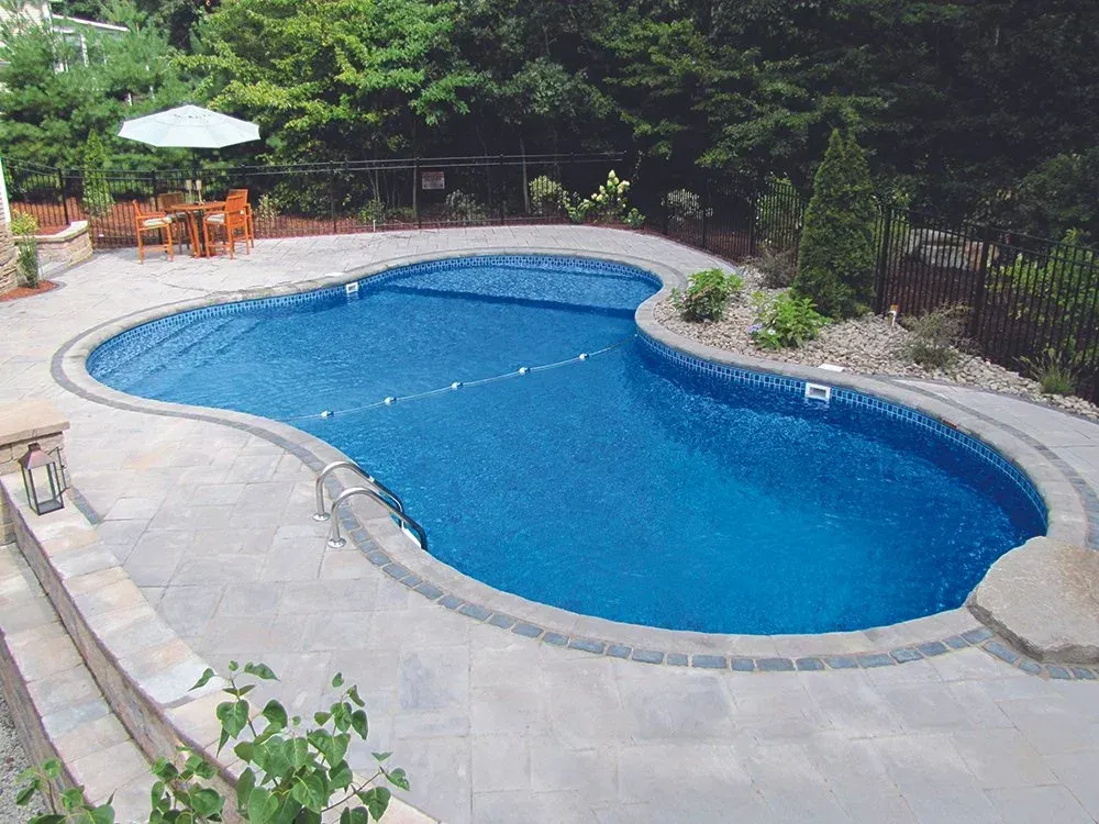 Pool with blue water and gray patio surrounded by greenery and seating.