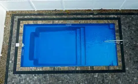 Overhead view of a rectangular blue swimming pool with steps, bordered by stone pavers and a white fence.