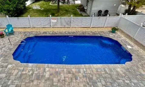 Blue swimming pool surrounded by a brick patio, fenced yard.