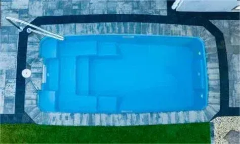 Aerial view of a rectangular blue swimming pool with stairs, surrounded by dark pavers and light-colored stonework.