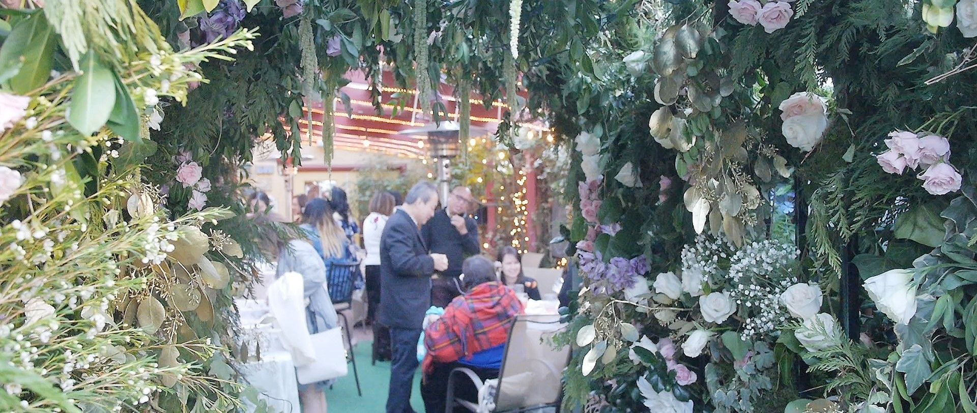 People walking through a flower-filled garden archway with hanging blossoms and lights