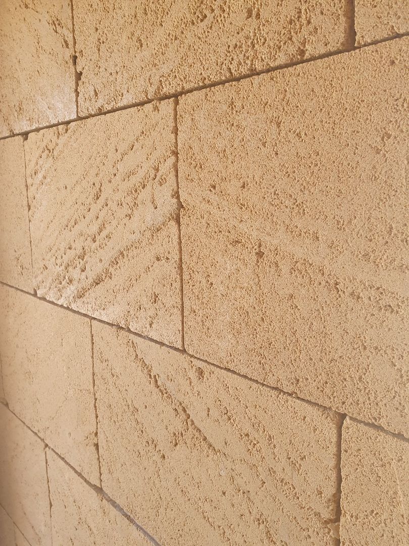 A textured wall made of rectangular, light-tan sandstone blocks with visible horizontal and vertical mortar joints.
