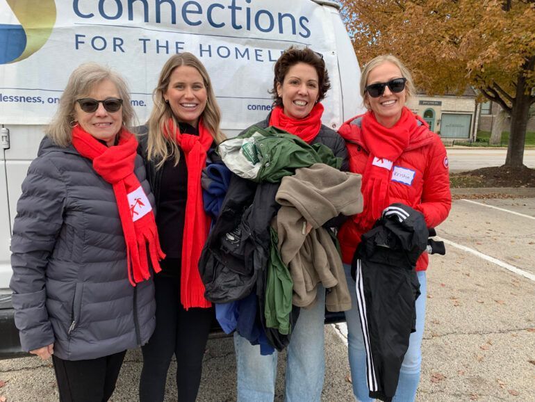 Four women wearing red scarves are standing in front of a van holding clothes.