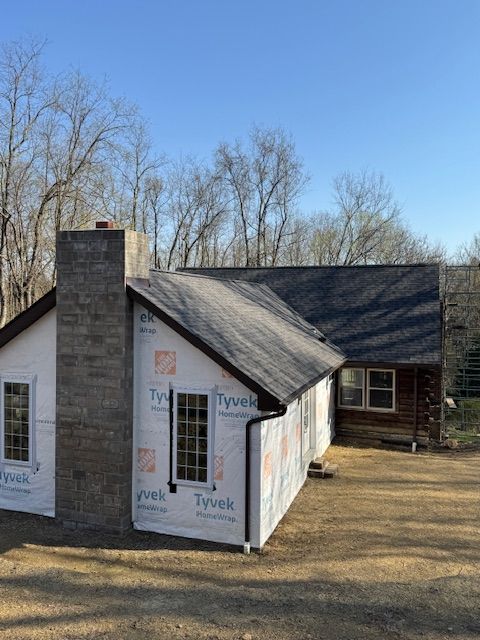 Construction of a house, with Tyvek wrap, stone chimney, dark roof, and windows, set against trees and a blue sky.