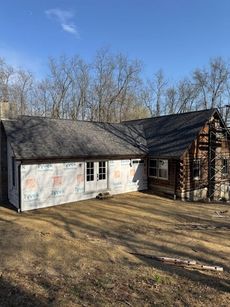 A log cabin under construction with new siding. White weather wrap covers the new frame, with a clear blue sky in the background.