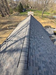 A gray asphalt shingle roof with a ridge cap runs from the foreground toward a chimney. A yard and trees are in the background.