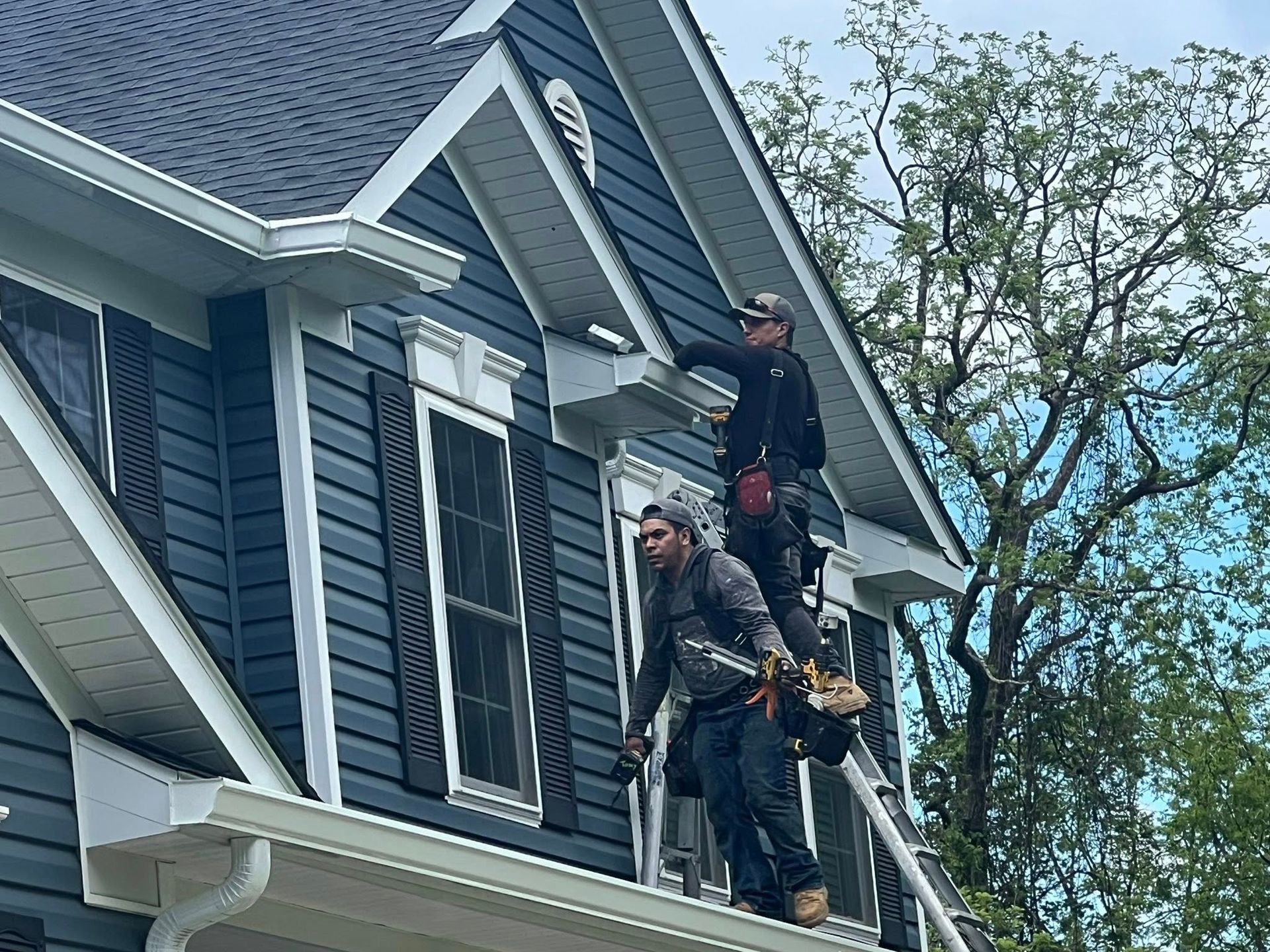 Two workers on a ladder installing something on a house's dark blue siding. The house has white trim and a dark roof.