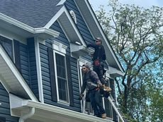 Two workers on a ladder installing something on a house's dark blue siding. The house has white trim and a dark roof.