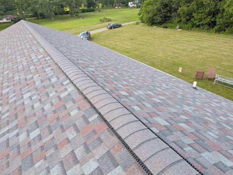 Aerial view of a roof with brown, grey, and red shingles. A central ridge runs the length of the roof. In the background is a grassy yard and trees.