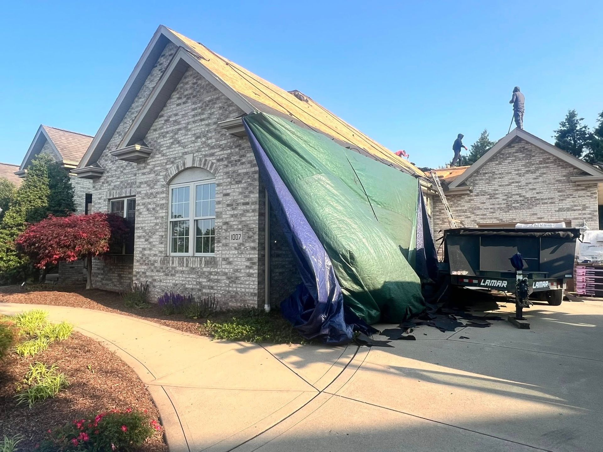 Home under roof repair with tarp covering the side; workers on the roof, dump truck, brick and landscaping.