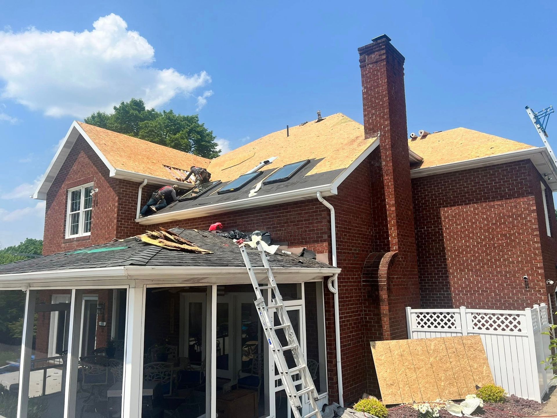 House with brick exterior, undergoing roof replacement; workers on roof, ladder against porch, blue sky.