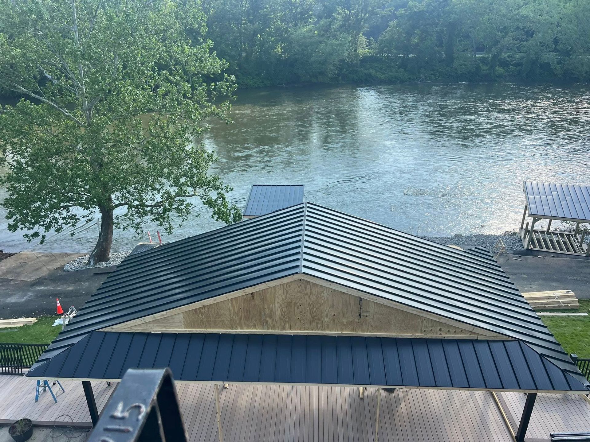 A dark blue metal roof with a water view. A tree and a river are in the background, and some construction materials are present.