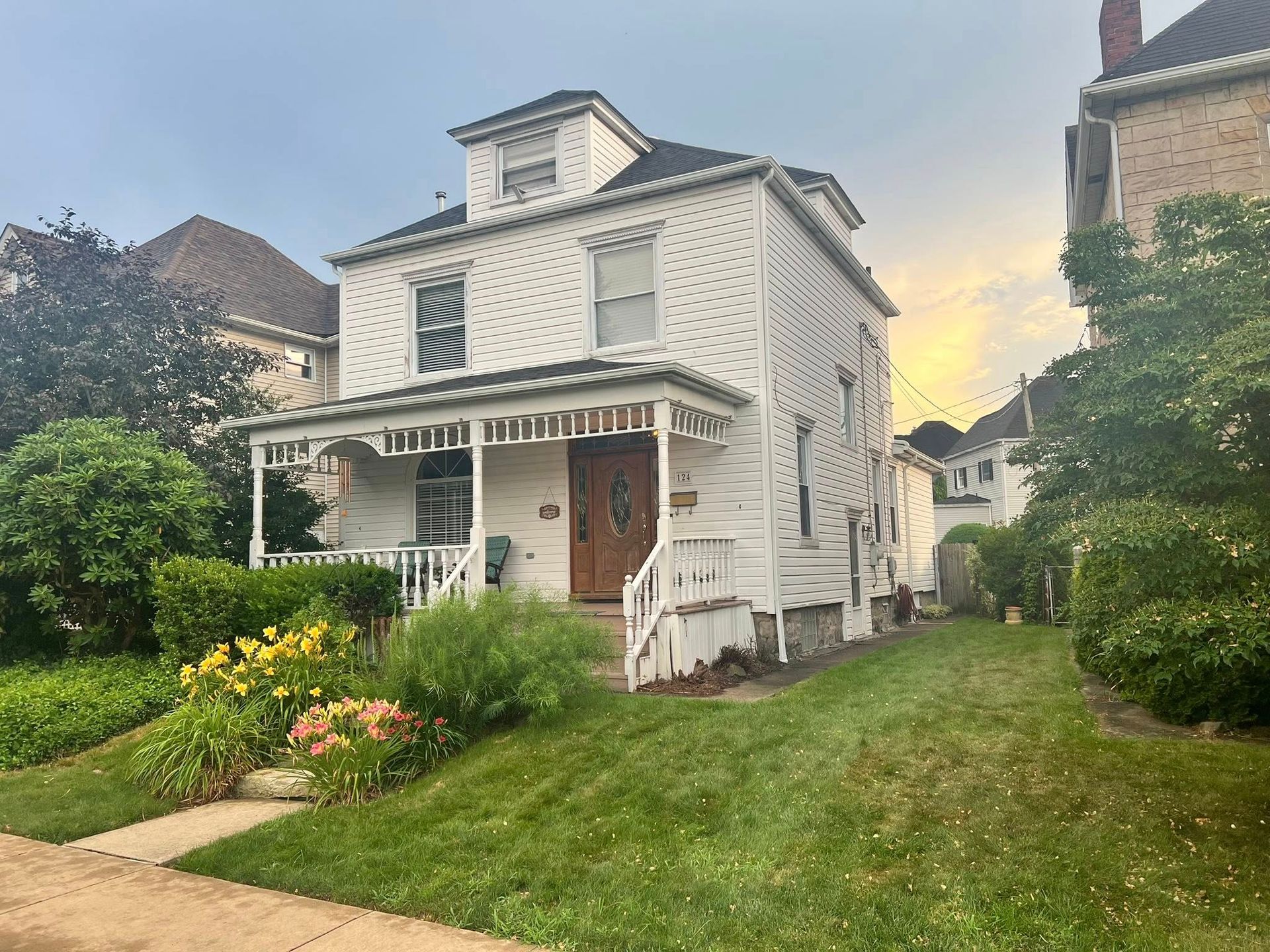 White two-story house with a porch and a brown door, set on a small green lawn with a few flowers and bushes.