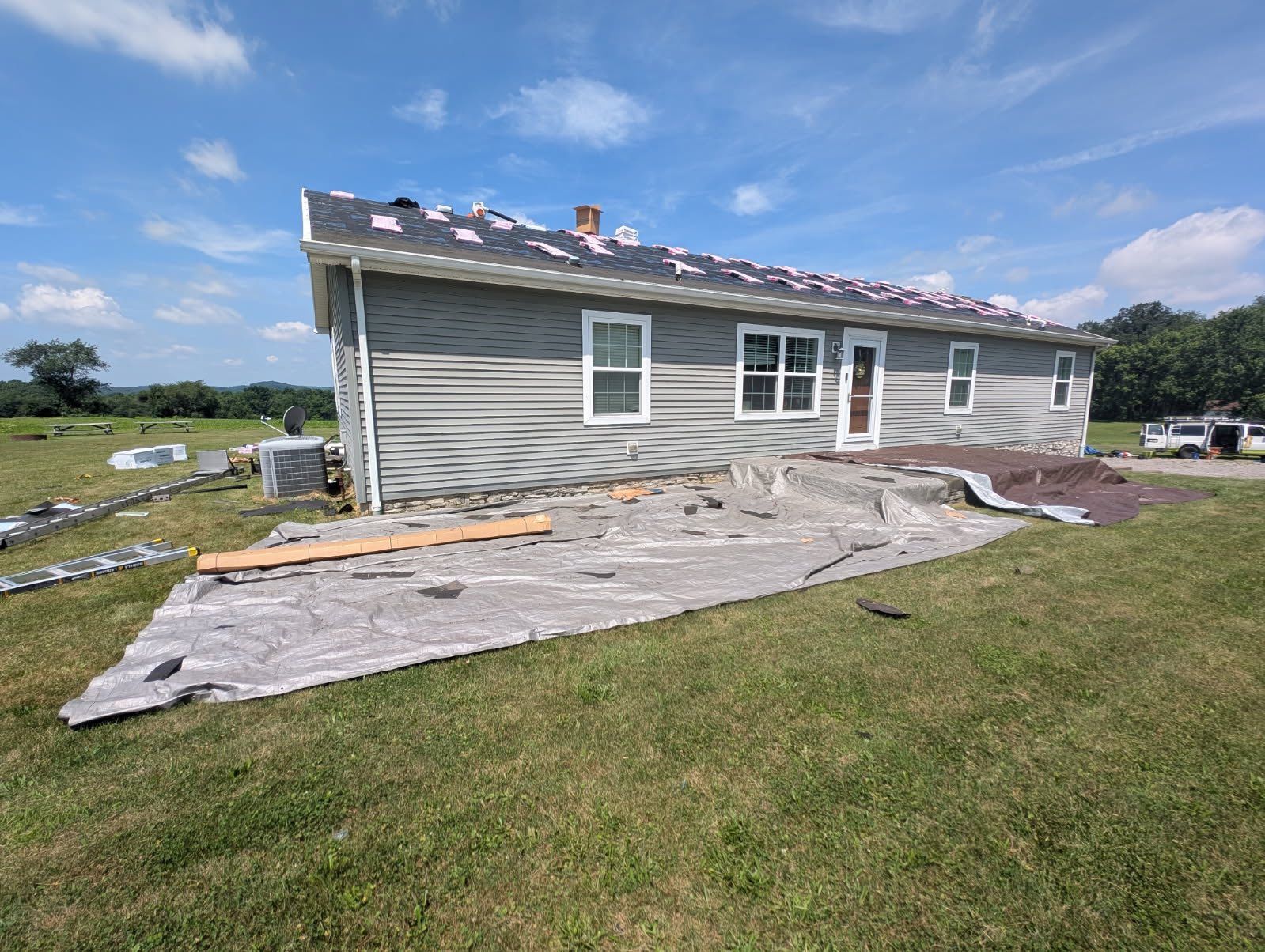 A house with gray siding and a partially replaced roof. Gray tarp and construction debris are in the yard.