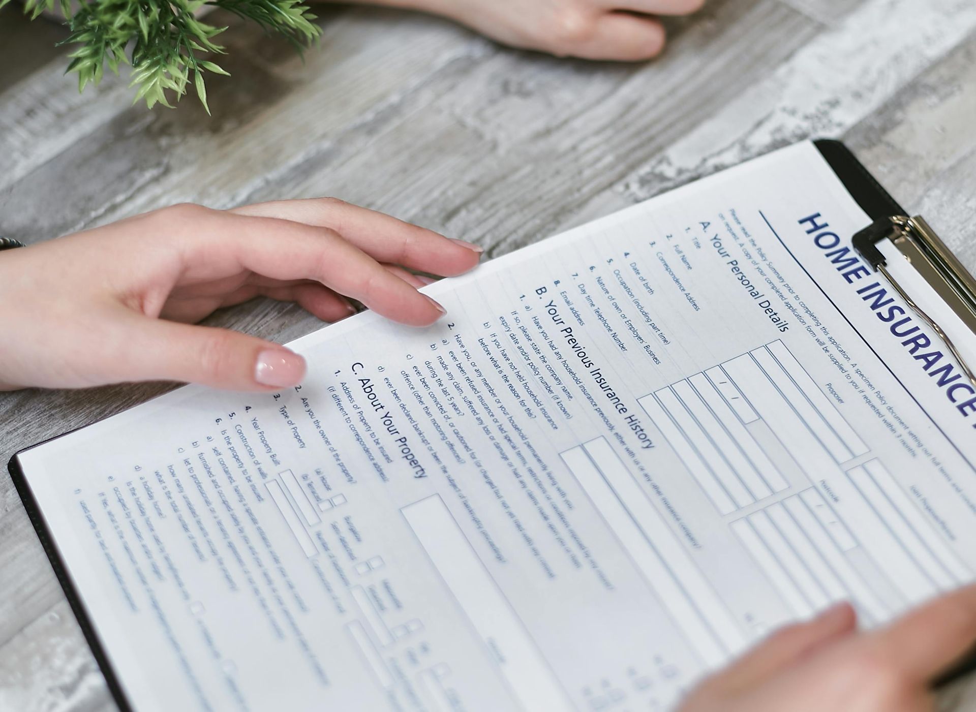 Hands filling out a home insurance form on a clipboard at a wooden table, likely at a desk; the form includes sections for personal details and property information.