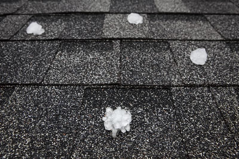 Hailstones rest on a dark gray asphalt shingle roof. The hailstones are white and irregularly shaped.