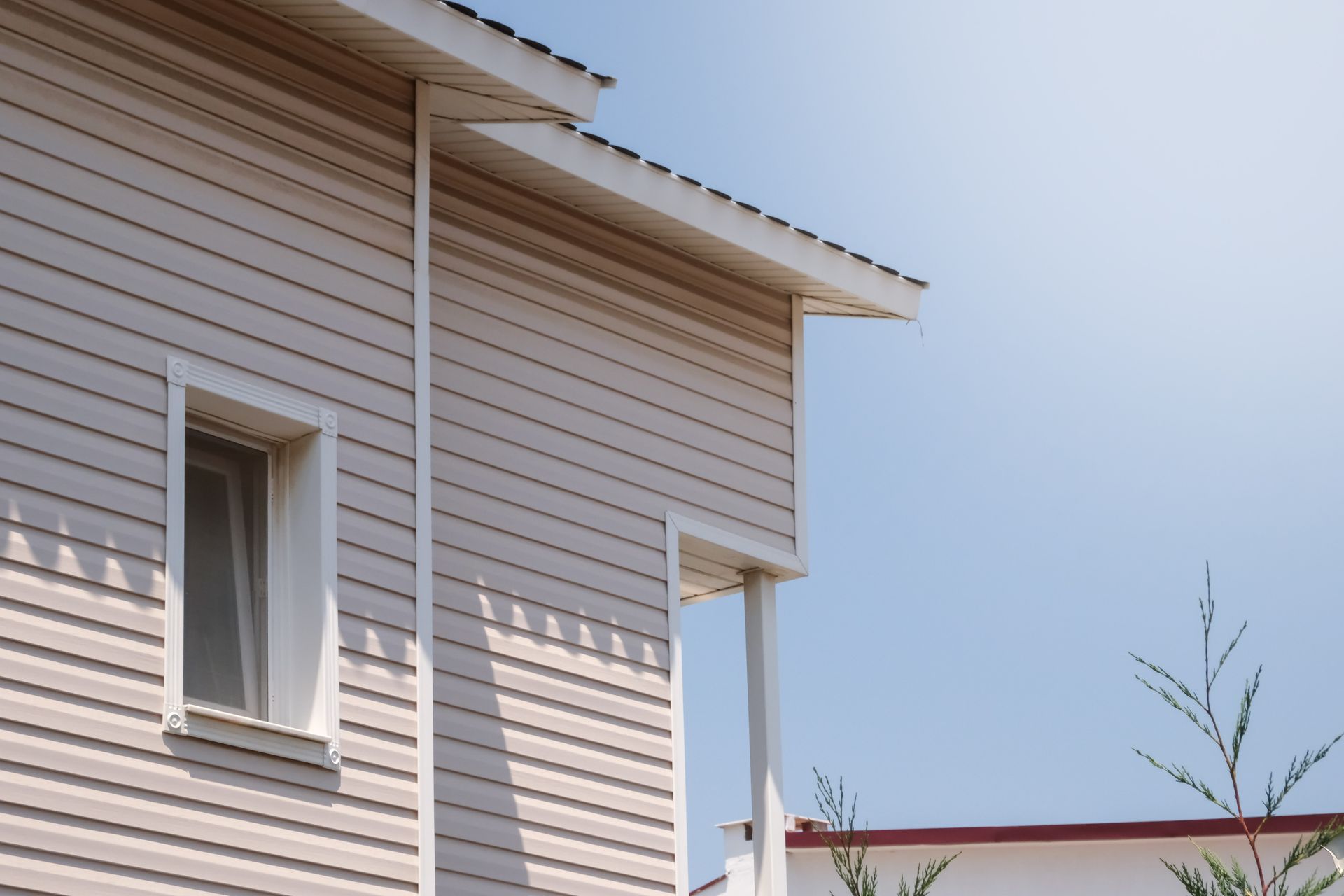 Beige vinyl-sided house with a window, white trim, and a blue sky background.