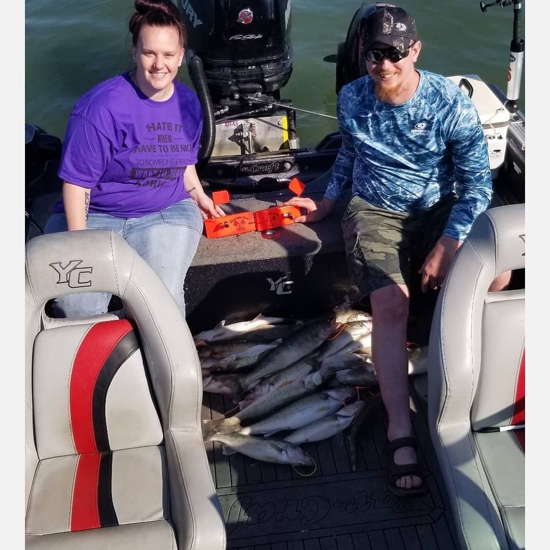 Two people on a boat with a pile of fish on the deck, smiling in bright sunlight.